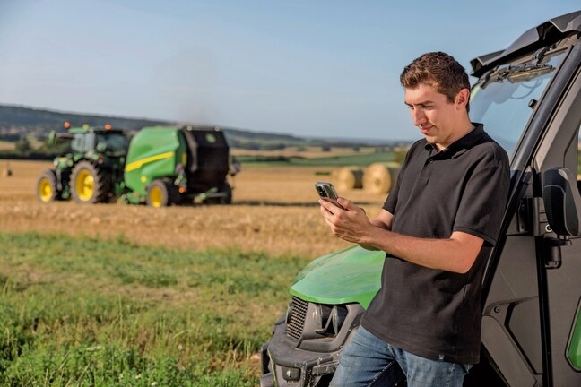 Person, der kontrollerer balledokumentationsdataene på mobiltelefonen med en John Deere VR-ballepresser i baggrunden