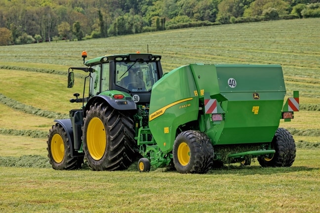 John Deere F442M ballepresser trukket af en John Deere 6M-traktor på en kuperet græsmark